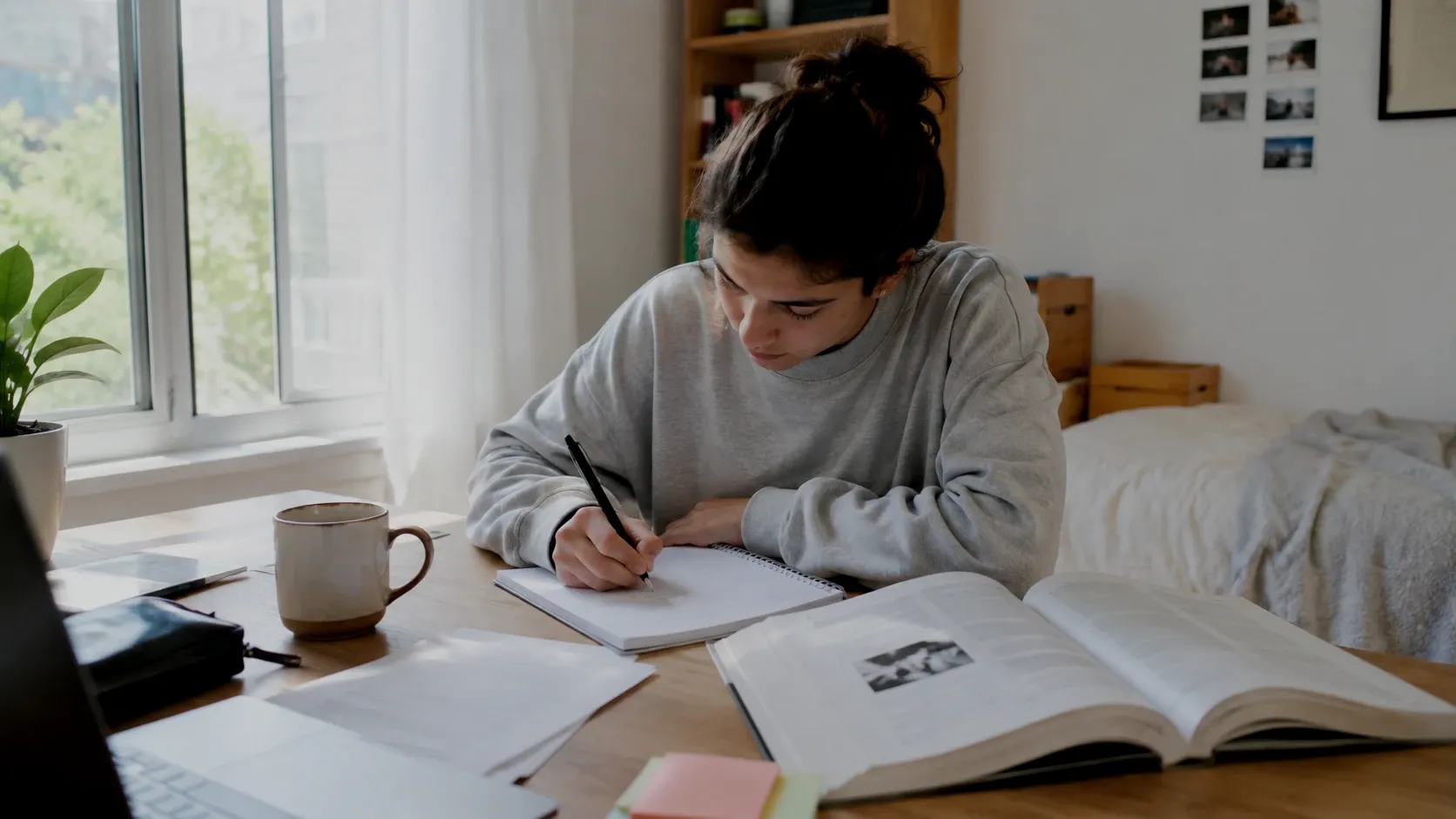 Persona estudiando en la mesa de su habitación, con apuntes y un libro abiertos junto a la ventana